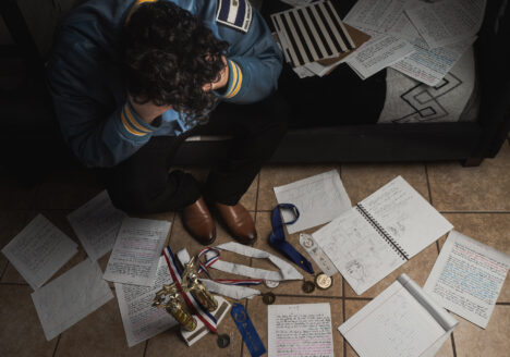 Photograph of young student sitting in their room with a mix of school papers, awards, and other items. They look distressed, and their face isn't in view.